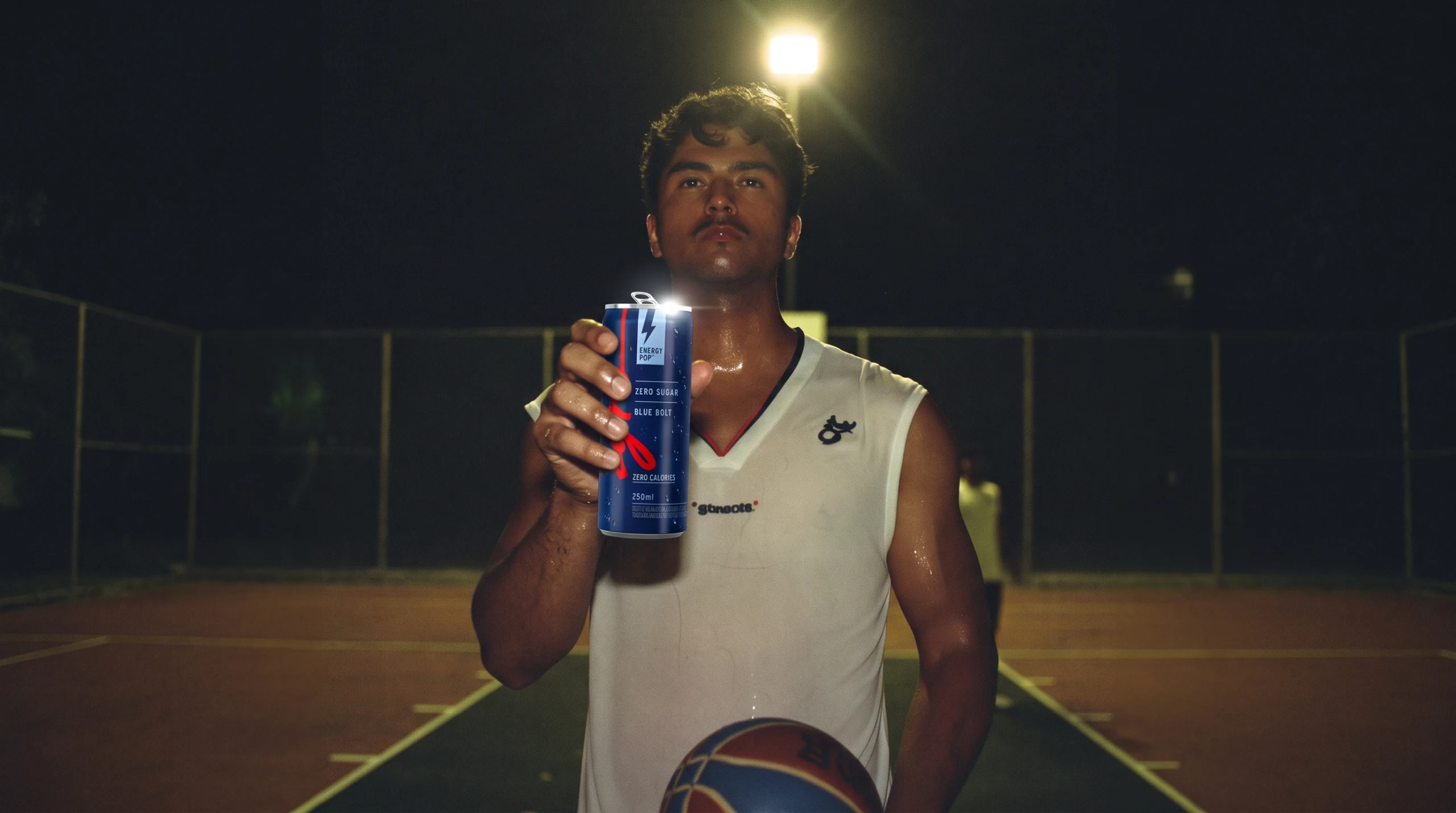 A basketball player holds up the blue can under a floodlight on a court at night.