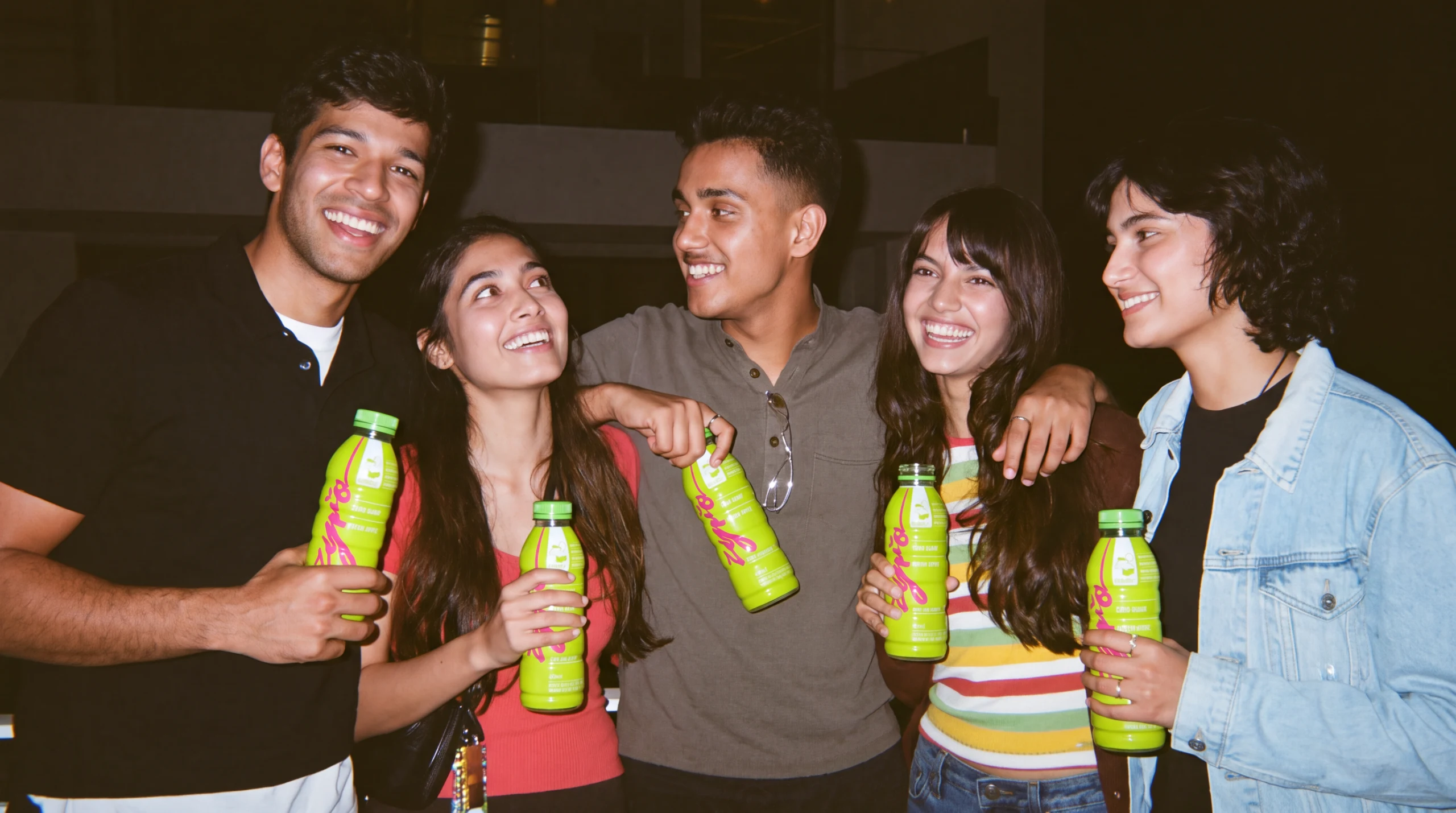Five friends laugh together at night, each holding a green apple bottle.