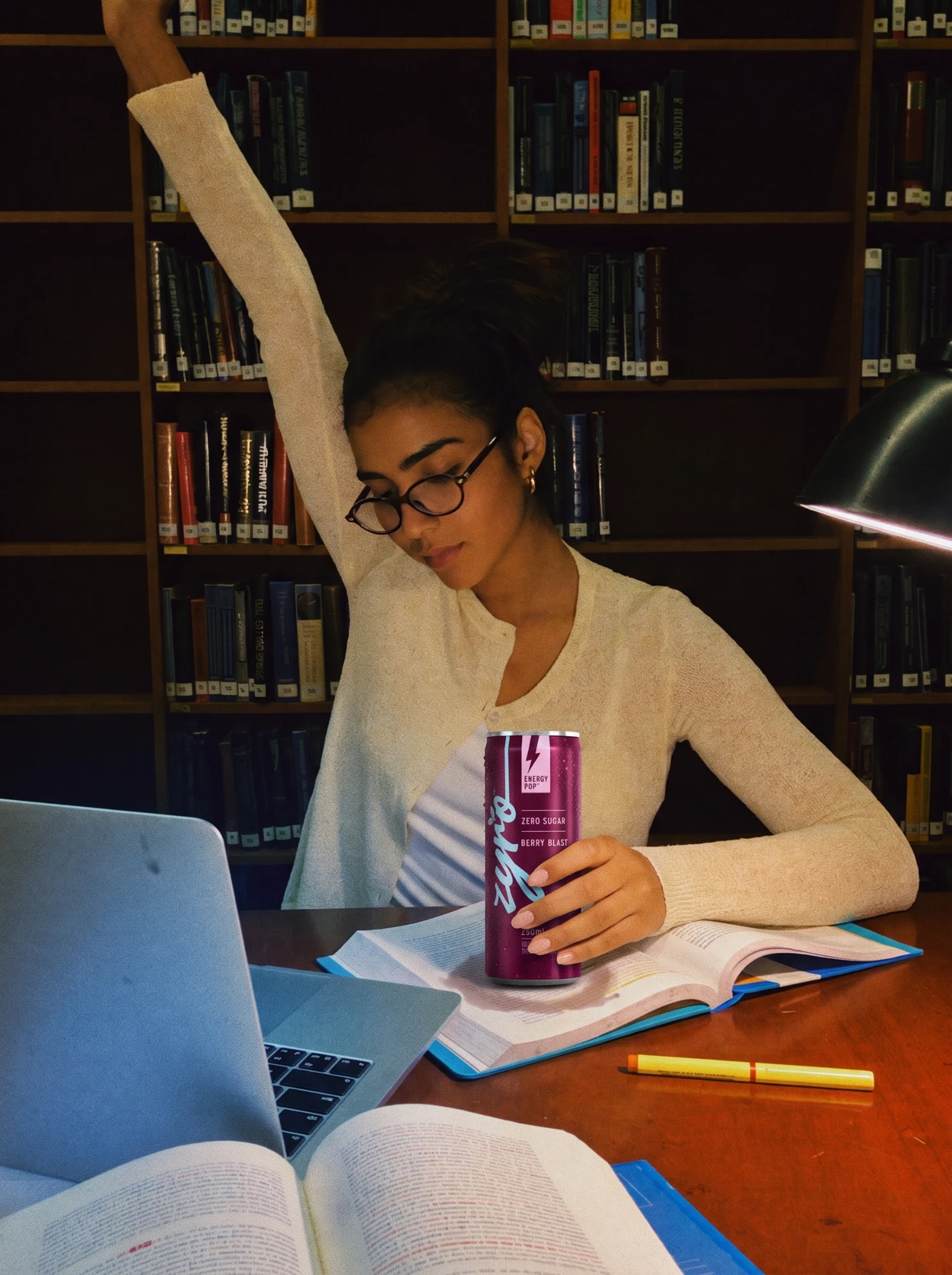 A student stretches at a library desk late at night, the berry can in her hand among open books and a laptop.