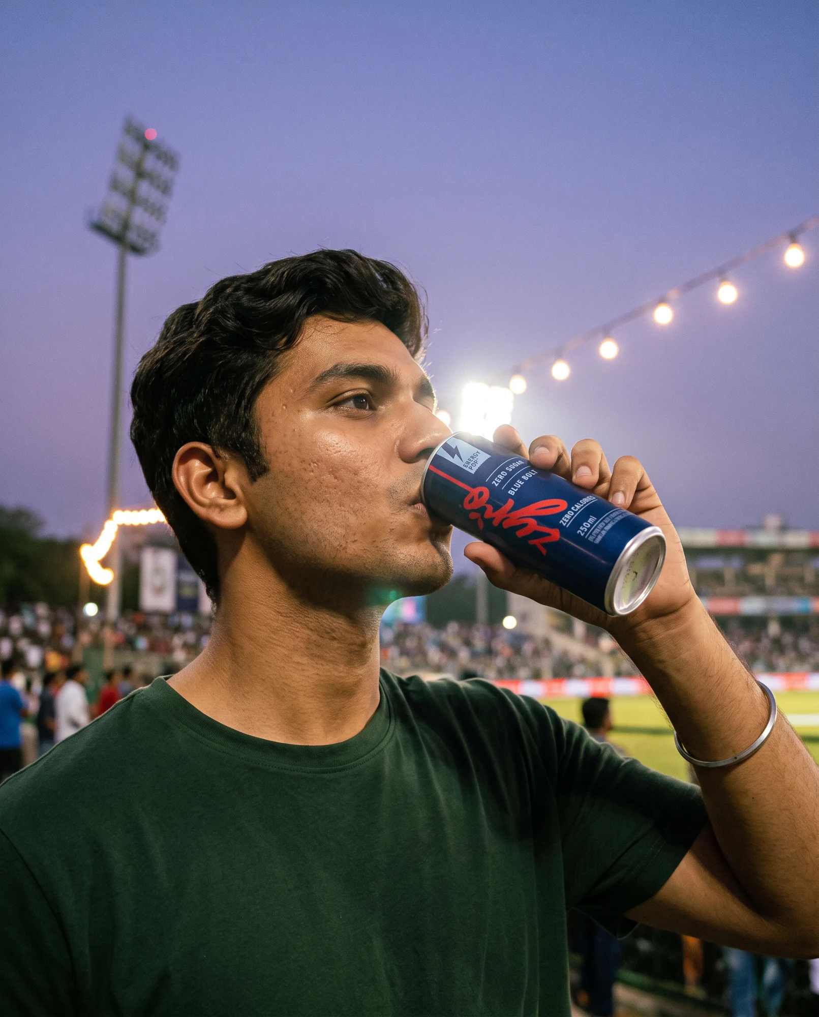 A fan drinks from the blue can in a cricket stadium at dusk, with floodlights and string lights behind.